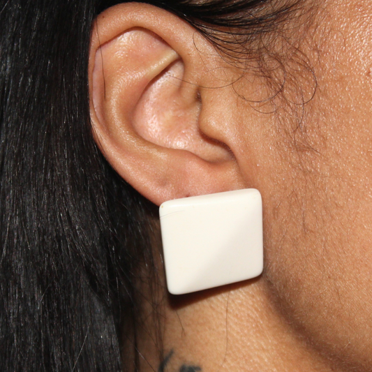 Close-up of an ear wearing a white square earring with dark hair and visible tattoo.